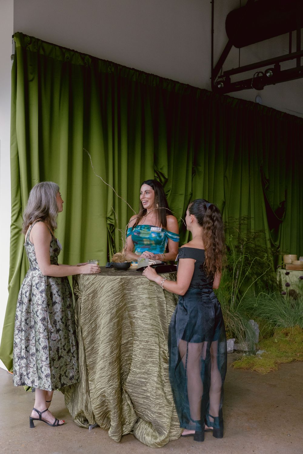 Three friends gather around a covered table with a green curtain backdrop in an event setting.