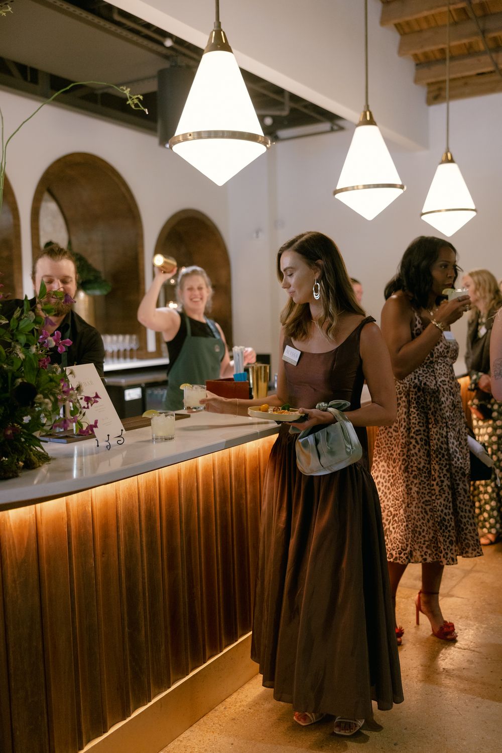 People gather at an illuminated wooden bar counter under pendant lights during a social event.