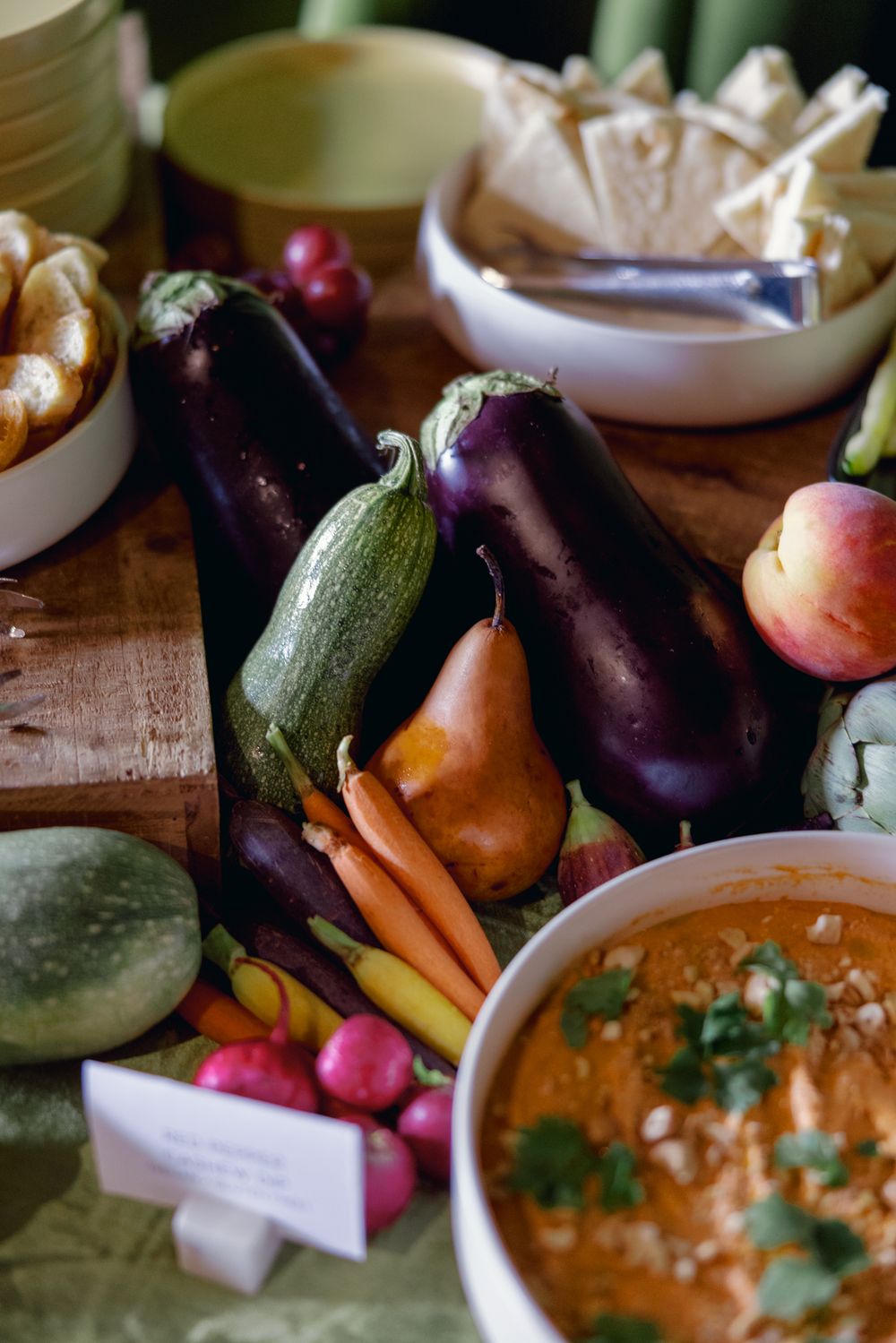 Fresh organic vegetables including eggplants, carrots, zucchini and radishes arranged on a rustic wooden surface with a bowl of dip.
