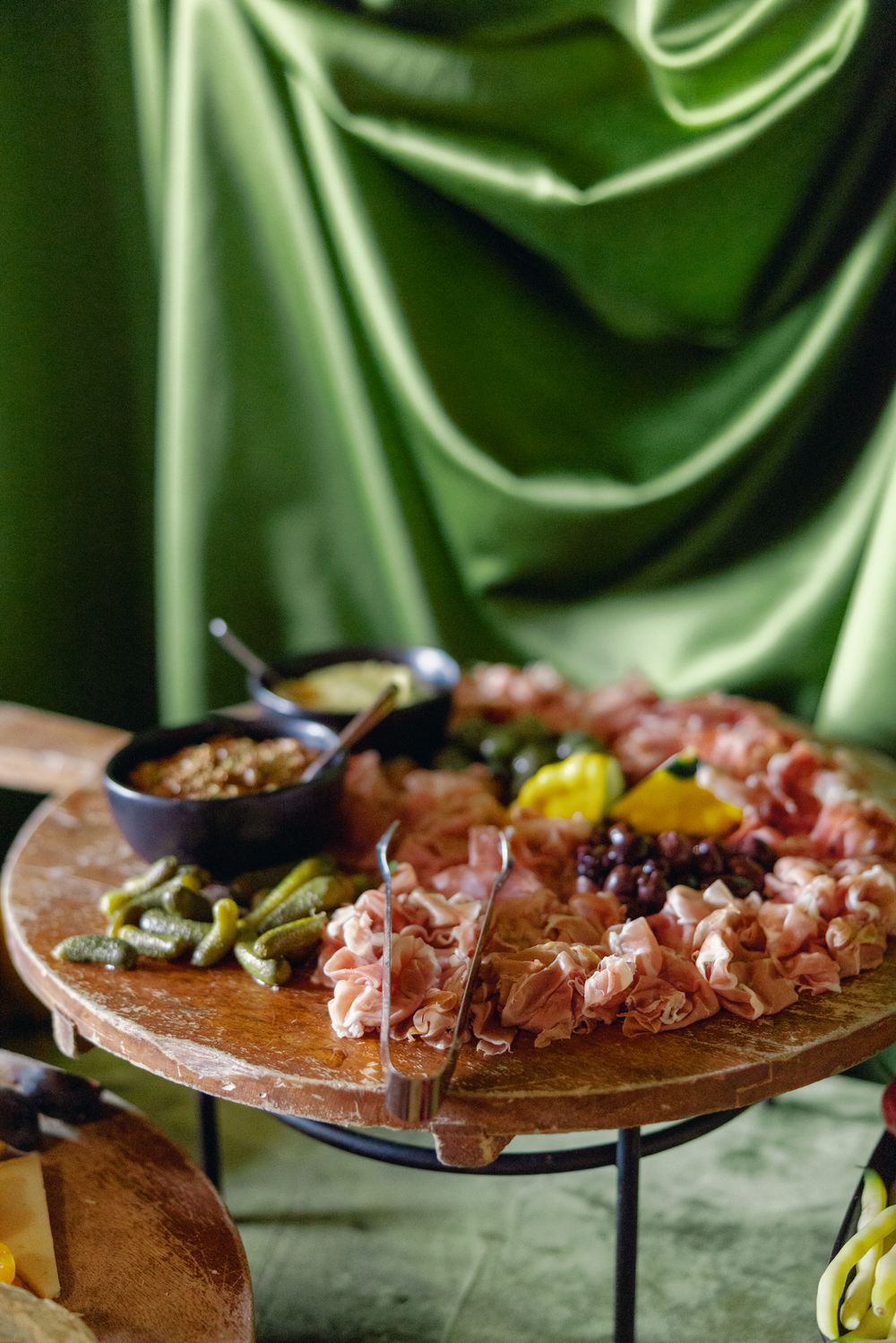 Charcuterie board with cured meats, olives, pickles and nuts displayed on wooden platter against green silk backdrop.