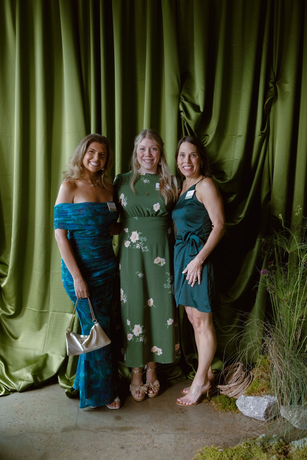 Three friends pose together against an emerald green curtain backdrop in formal dresses for a special occasion.
