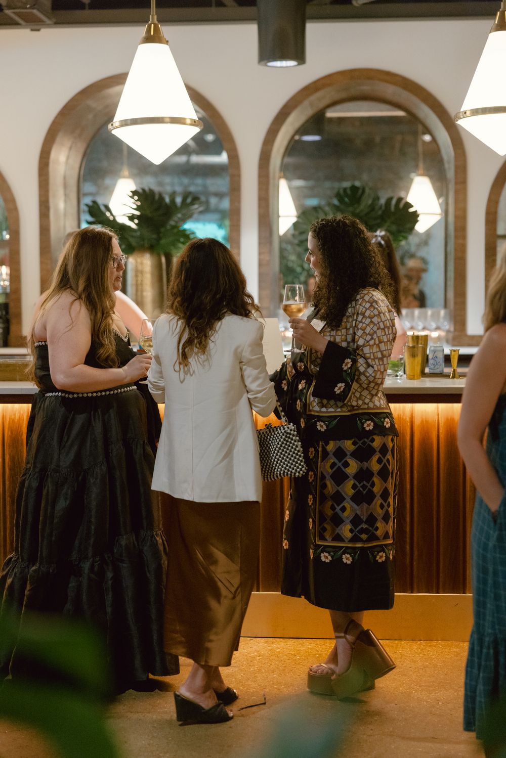 Women chatting by an illuminated bar with arched mirrors and pendant lighting at an elegant venue.