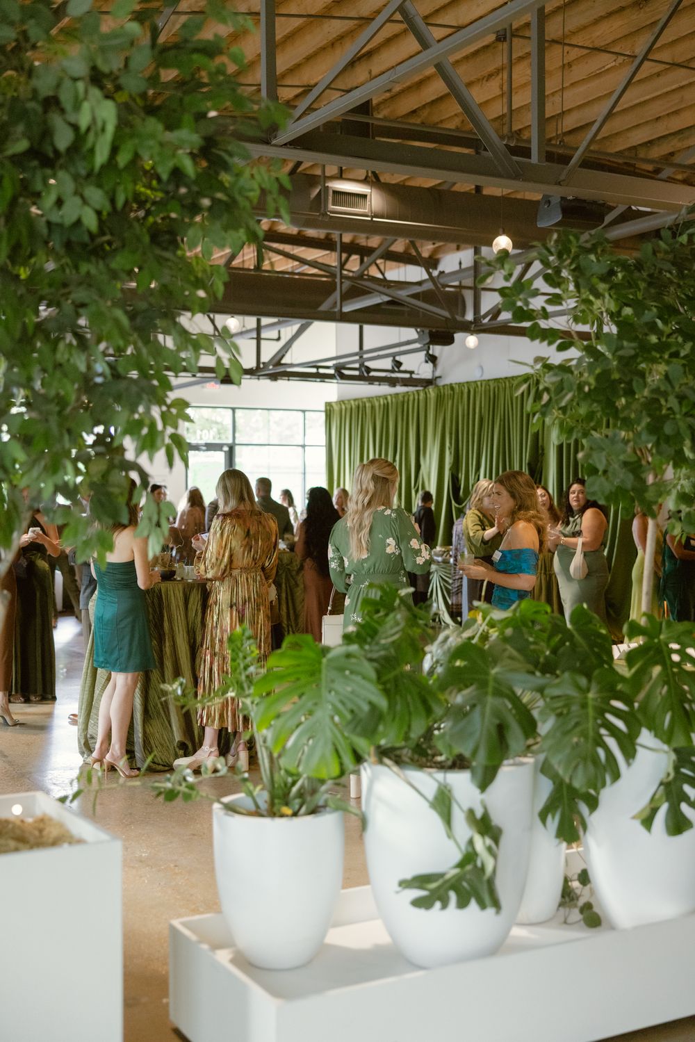 Stylishly dressed event guests socialize among large potted monstera plants in an industrial-chic space.