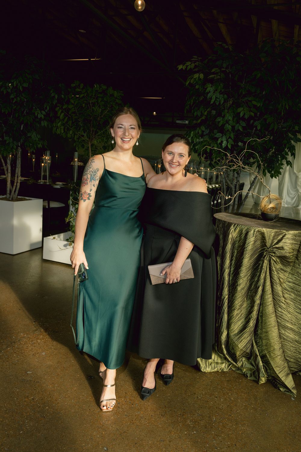 Two people in elegant formal dresses pose together at an evening event with green decor.