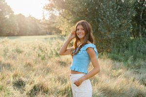 Woman in light blue top stands in golden field at sunset with trees in background.