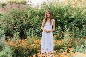 A woman in a white dress stands surrounded by colorful wildflowers and lush greenery in a garden setting.