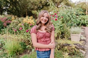 Smiling teenage girl in pink top and jeans posing in a lush garden with colorful roses blooming behind her.