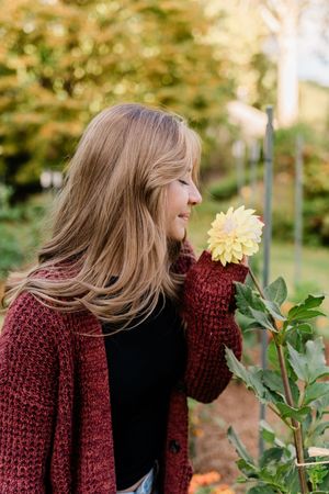 Young woman in red cardigan smelling a yellow dahlia flower in an autumn garden.
