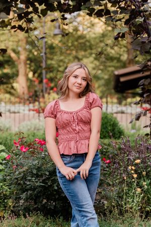 Young woman in pink top and jeans poses among colorful garden roses and lush greenery.