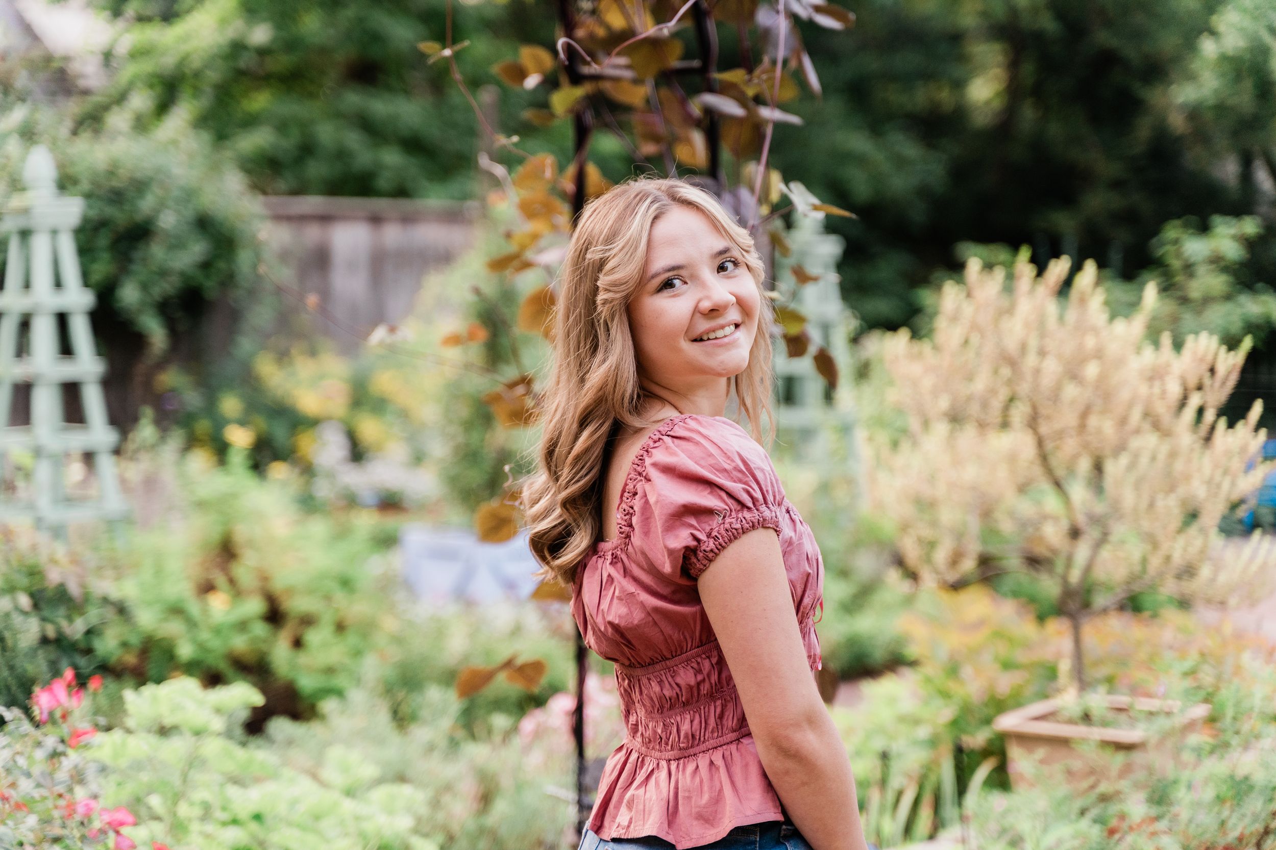 Smiling blonde woman in pink top standing in lush garden surrounded by green plants and colorful foliage.