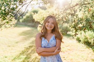 Happy teenage girl in blue floral dress standing under sunlit tree branches with arms crossed.