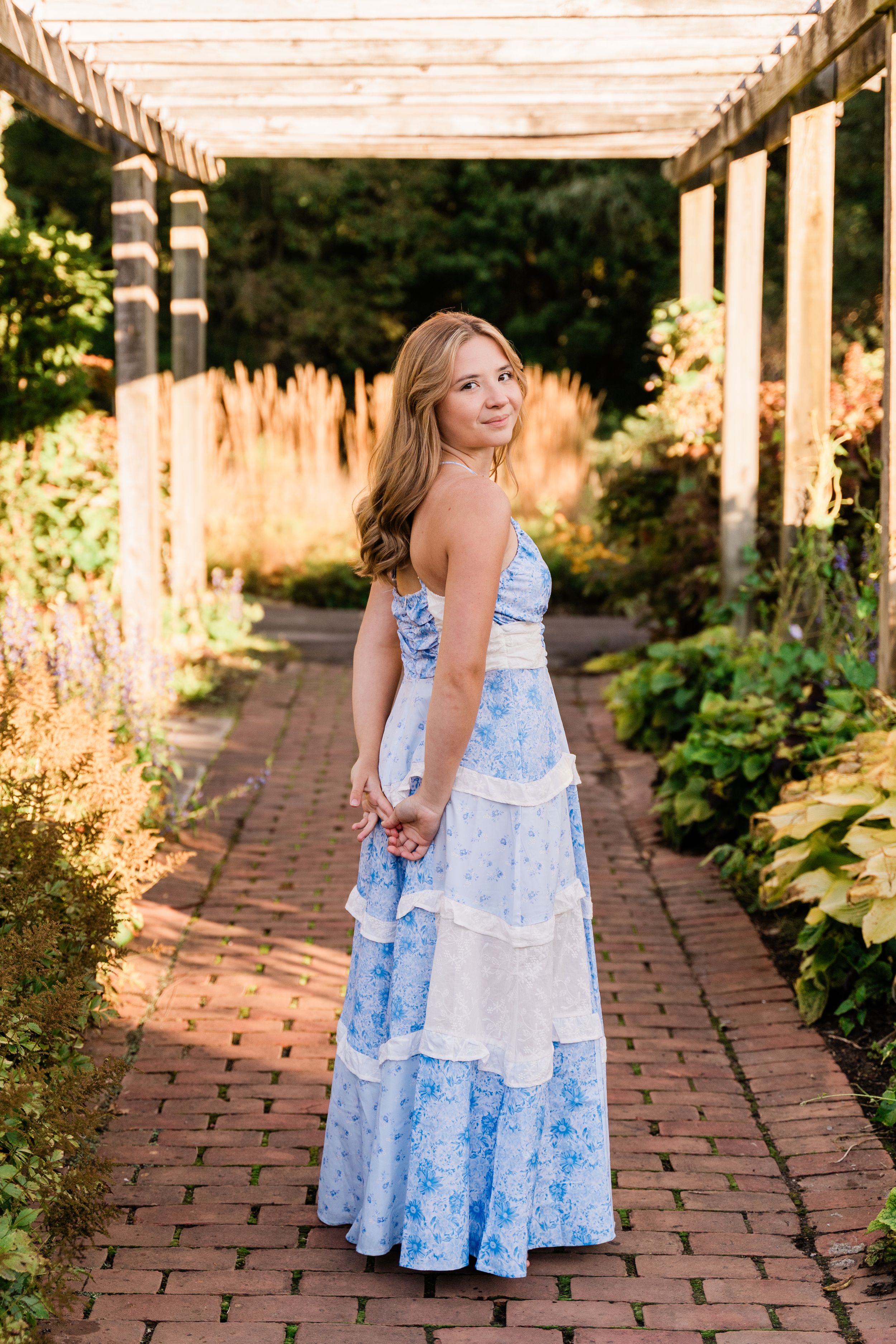 Young woman in blue floral maxi dress posing on brick path under garden pergola.