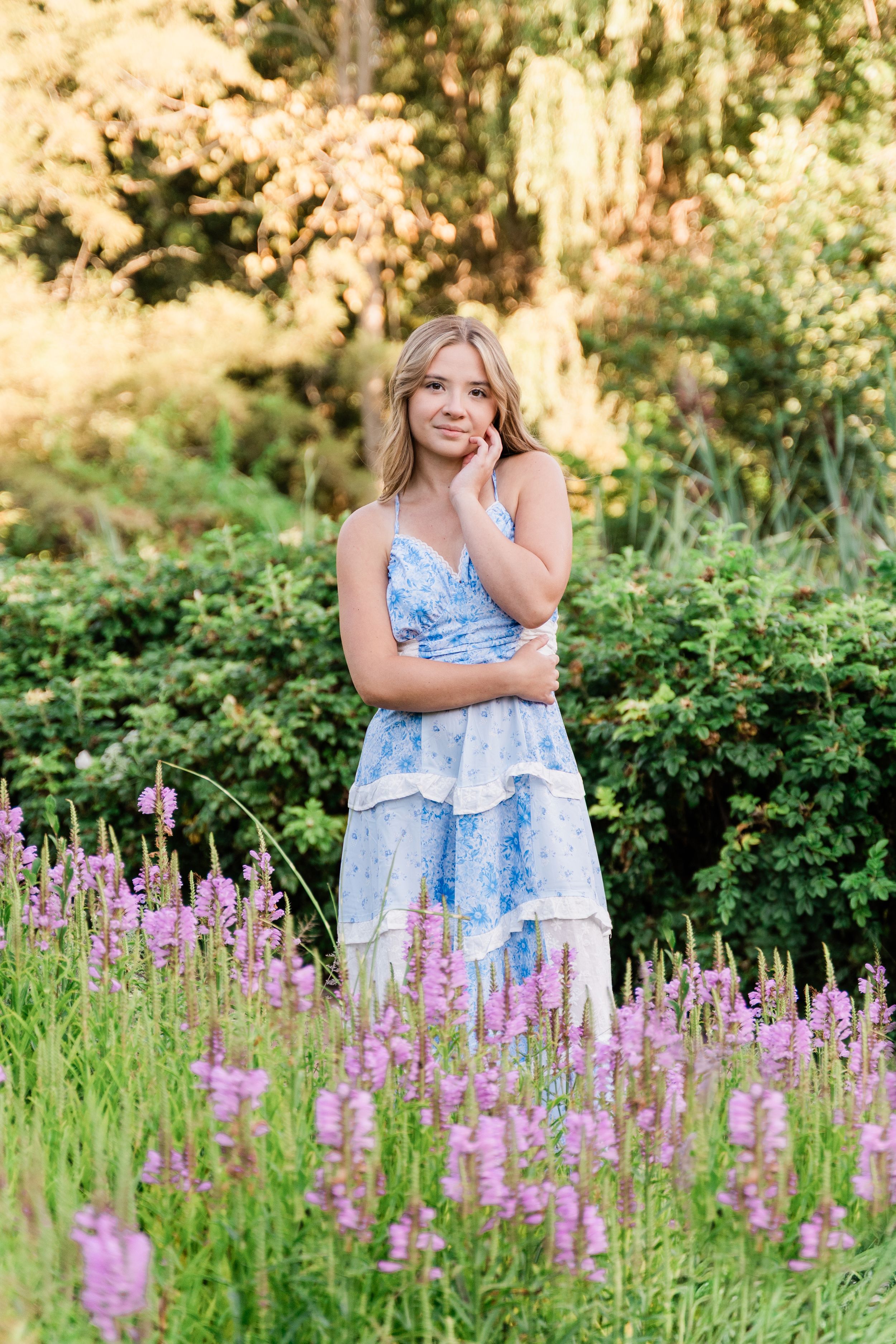 Blonde woman in blue floral dress standing among purple wildflowers in garden.