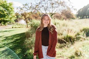 Smiling young woman in red cardigan and black top stands in sunny park setting.