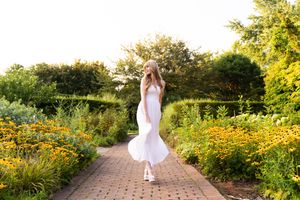 Person in flowing white dress walks along brick path surrounded by vibrant summer garden flowers and greenery.