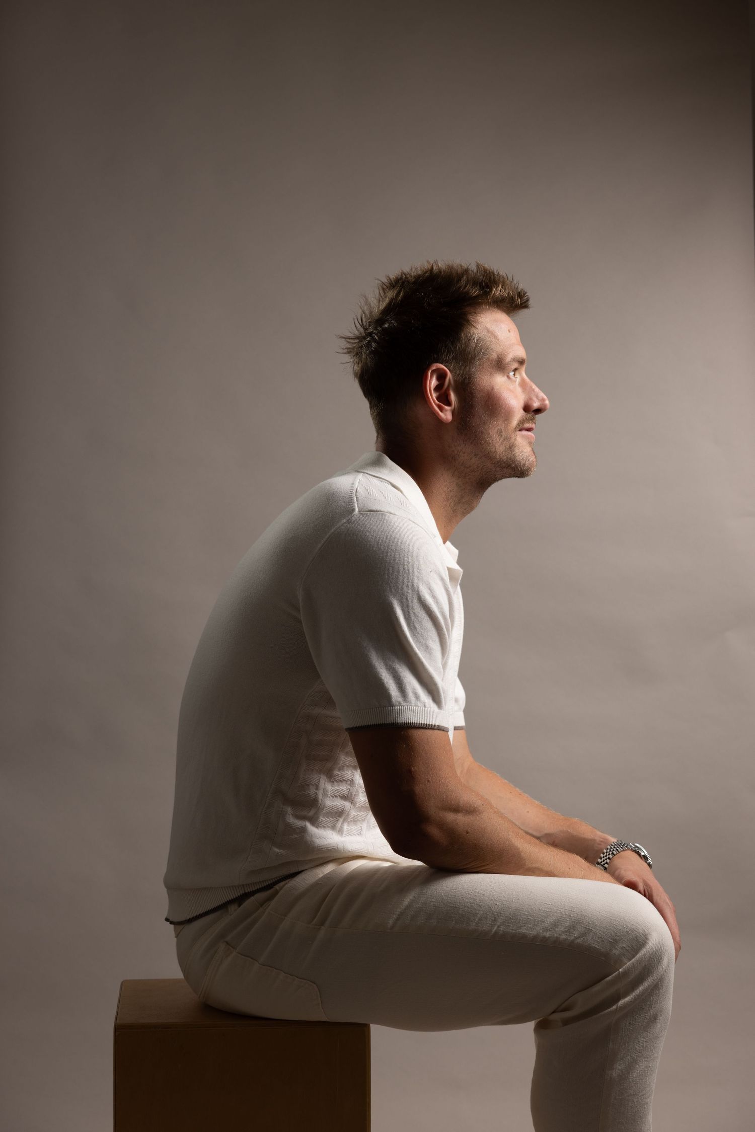 Profile view of person in all white clothing sitting on brown cube against neutral gray background in studio setting.