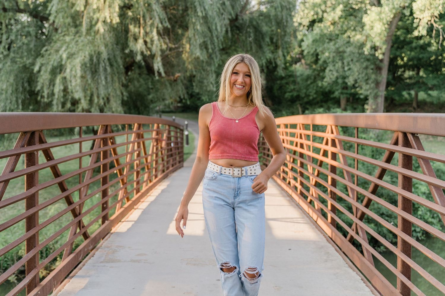 Toledo area senior poses on a rustic red bridge in a lush natural setting while wearing a pink tank top and ripped jeans.