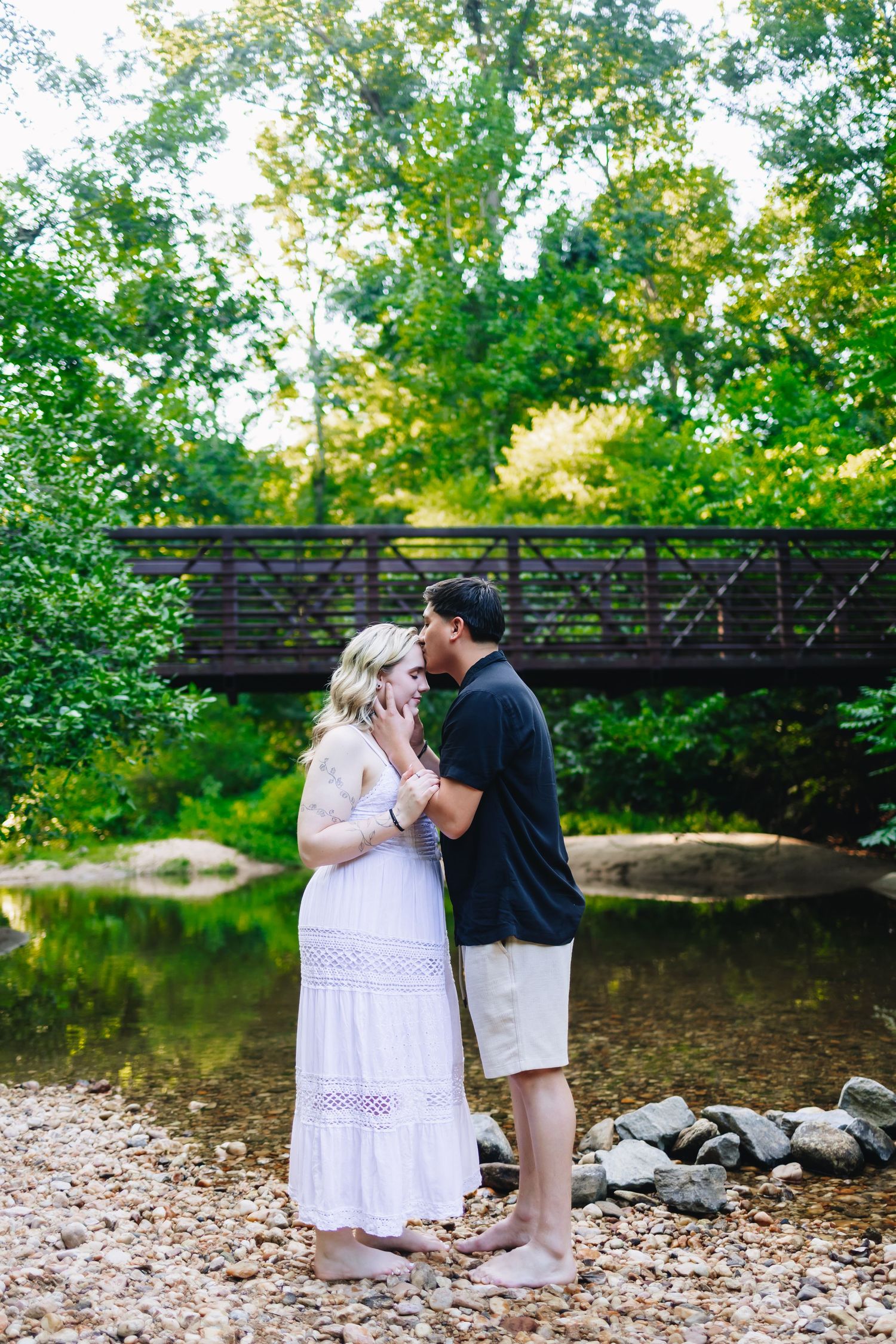 Couple embraces near footbridge with green foliage and stream in natural outdoor setting.