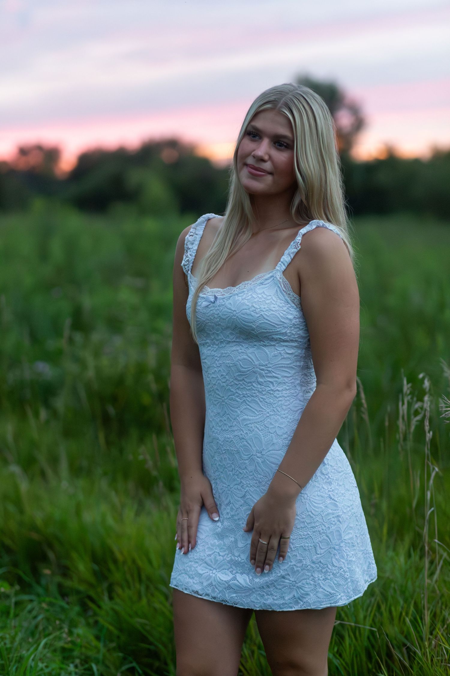 Woman in white lace top poses in green field at sunset.