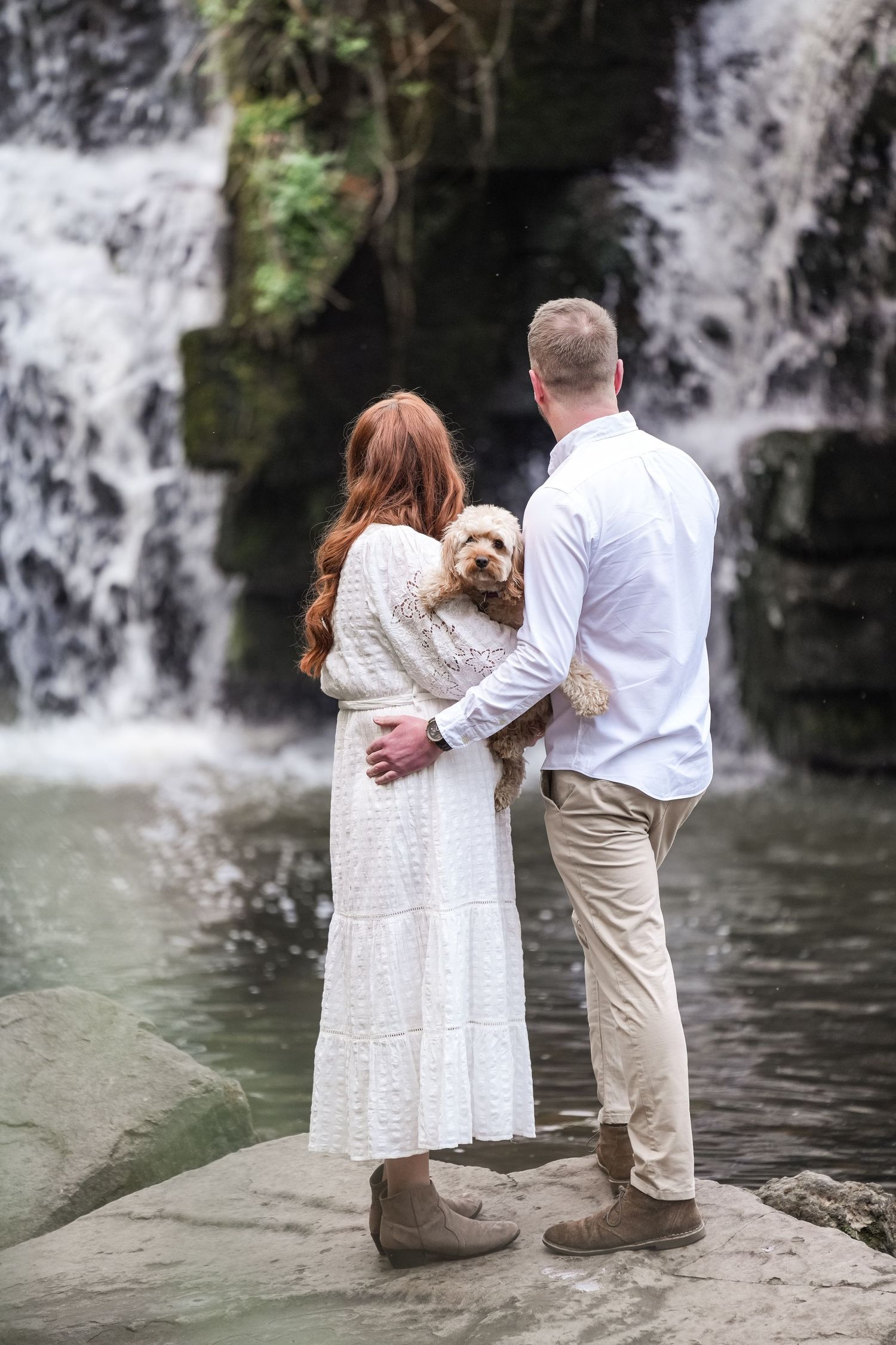 A couple stands with their dog on a rock overlooking a beautiful cascading waterfall in a serene natural setting.
Engagement photoshoot at