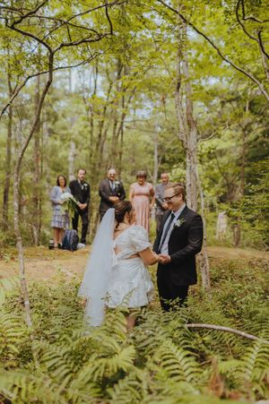 A wedding ceremony takes place in a lush forest setting with ferns and green foliage surrounding the couple.