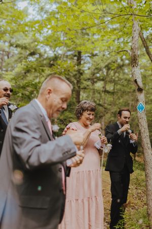 Wedding guests in formal attire gather under trees during an outdoor ceremony.