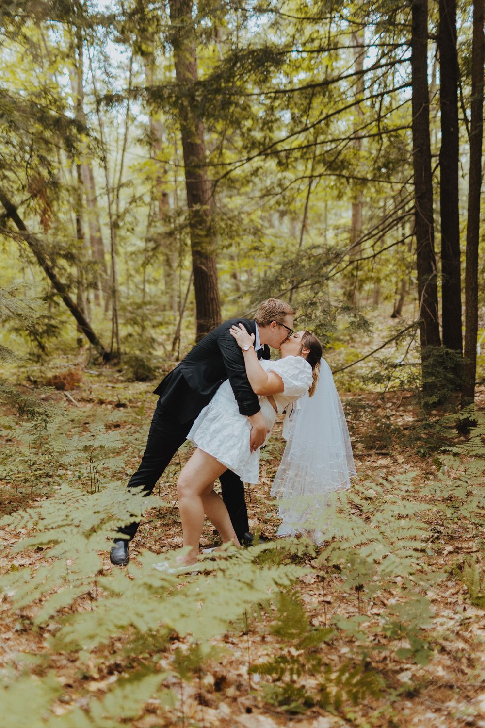 A romantic dip kiss between newlyweds in a sunlit forest setting.