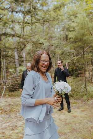 A guest in a blue dress holds a white bouquet during an outdoor forest wedding.