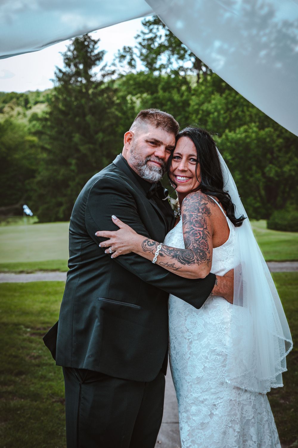 A couple embraces under a white wedding canopy during their outdoor ceremony on a green lawn.