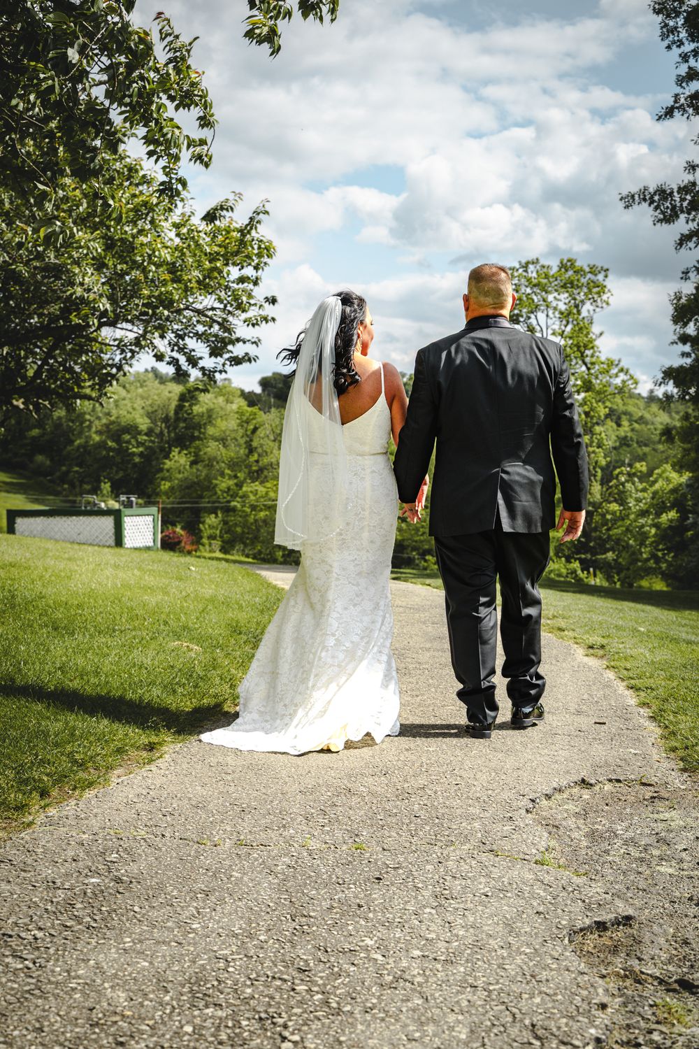 A couple in wedding attire walking along a scenic path surrounded by green trees and grass on a cloudy day.