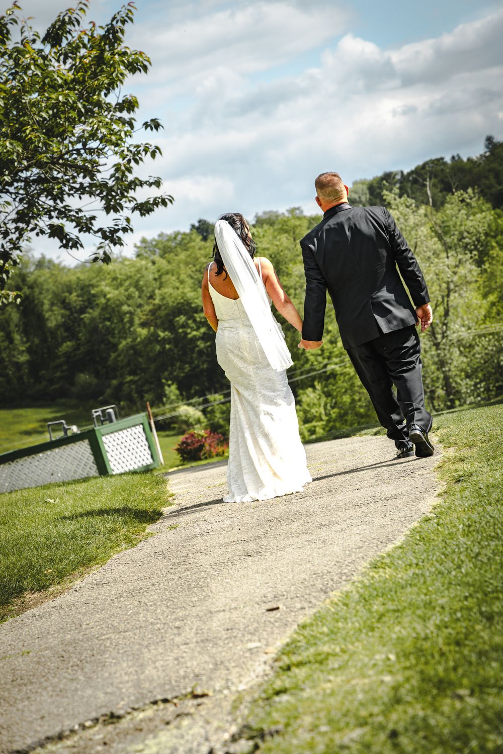 A couple walking along a garden path on their wedding day, with the bride in white and groom in black suit.