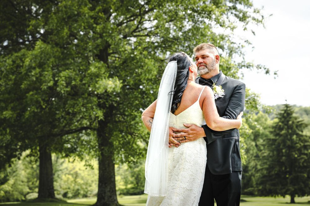 Couple embraces during outdoor wedding ceremony in a serene park setting with lush green trees in the background.