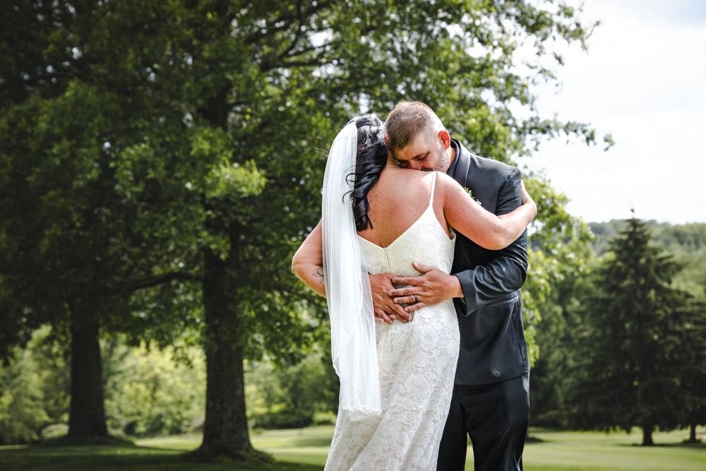 Newlywed couple embraces outdoors on golf course with trees in background during summer wedding ceremony.
