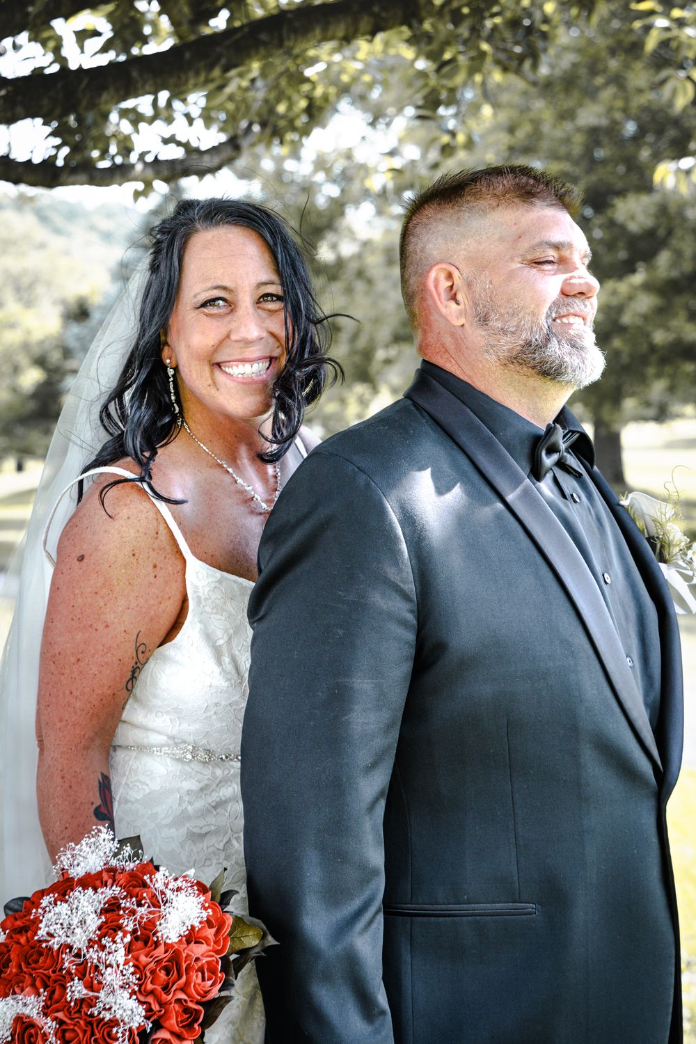 Happy couple laughing together in formal wedding attire with vibrant red rose bouquet outdoors.