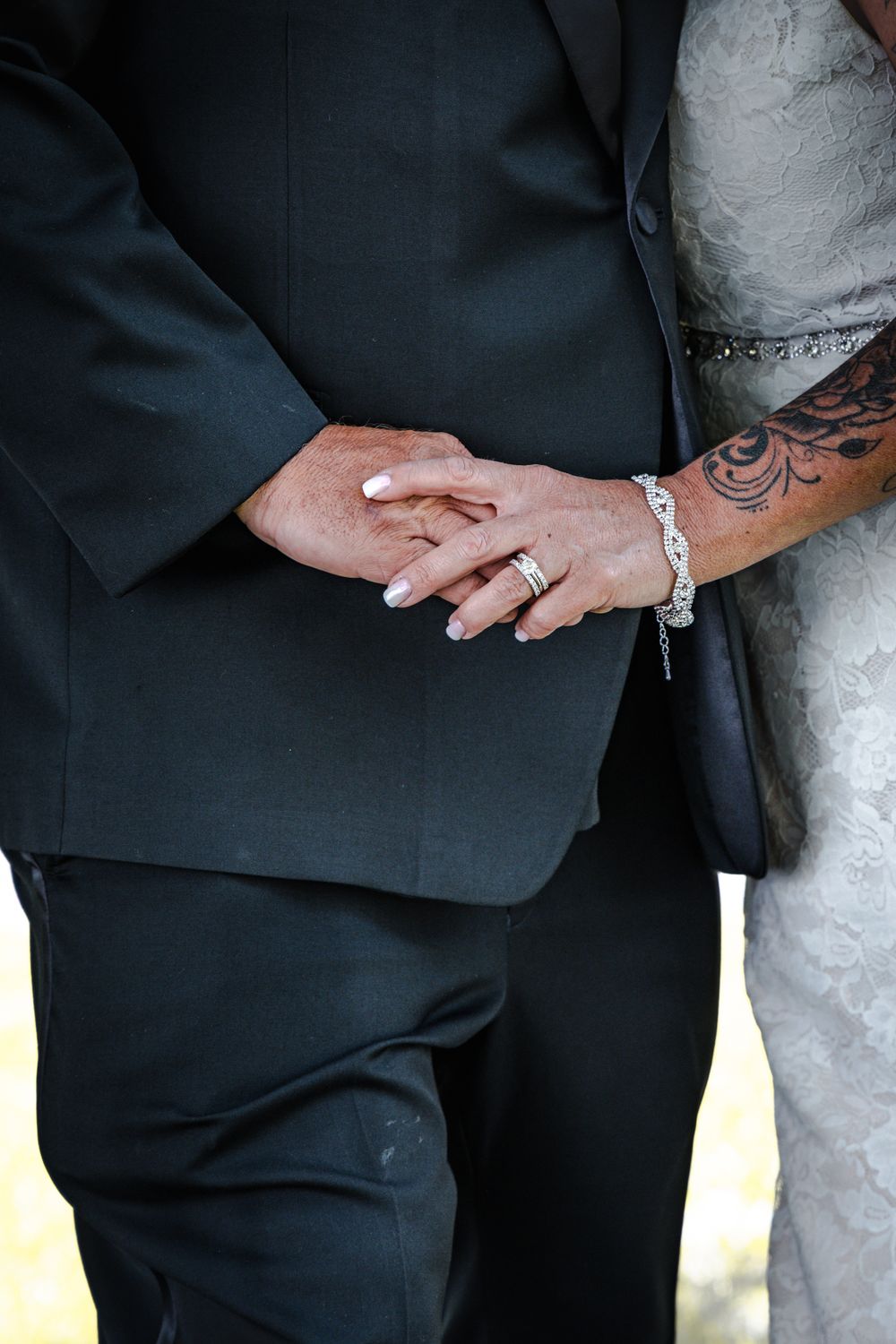 Close up of newlywed couple's hands showing wedding rings and a tattoo against formal attire.