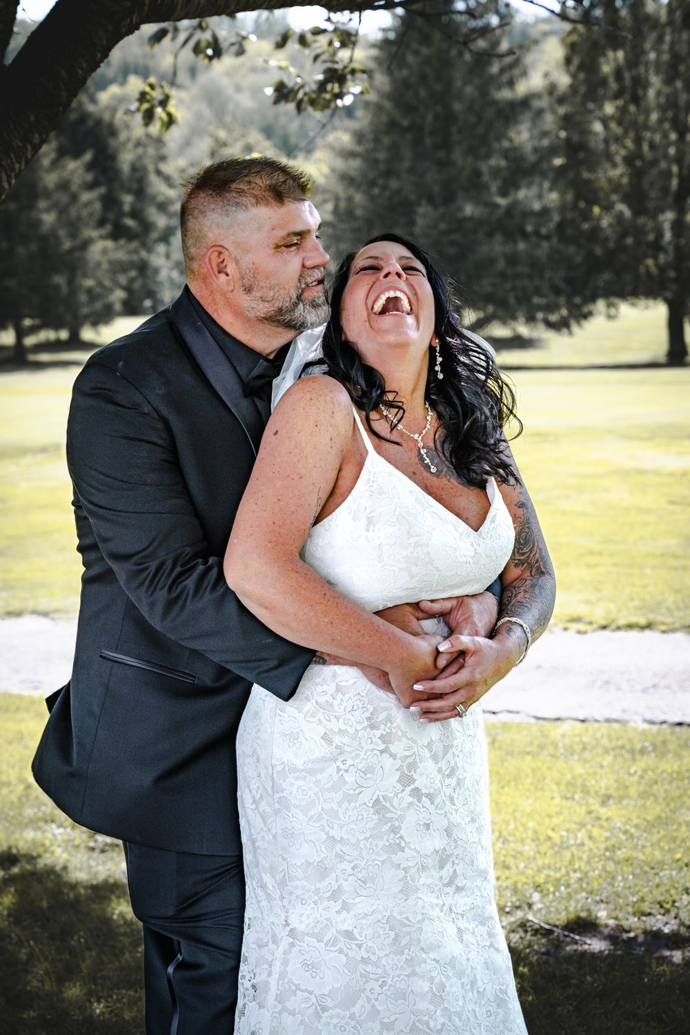 A joyful couple shares a candid moment of laughter in their wedding attire on a sunny day outdoors.