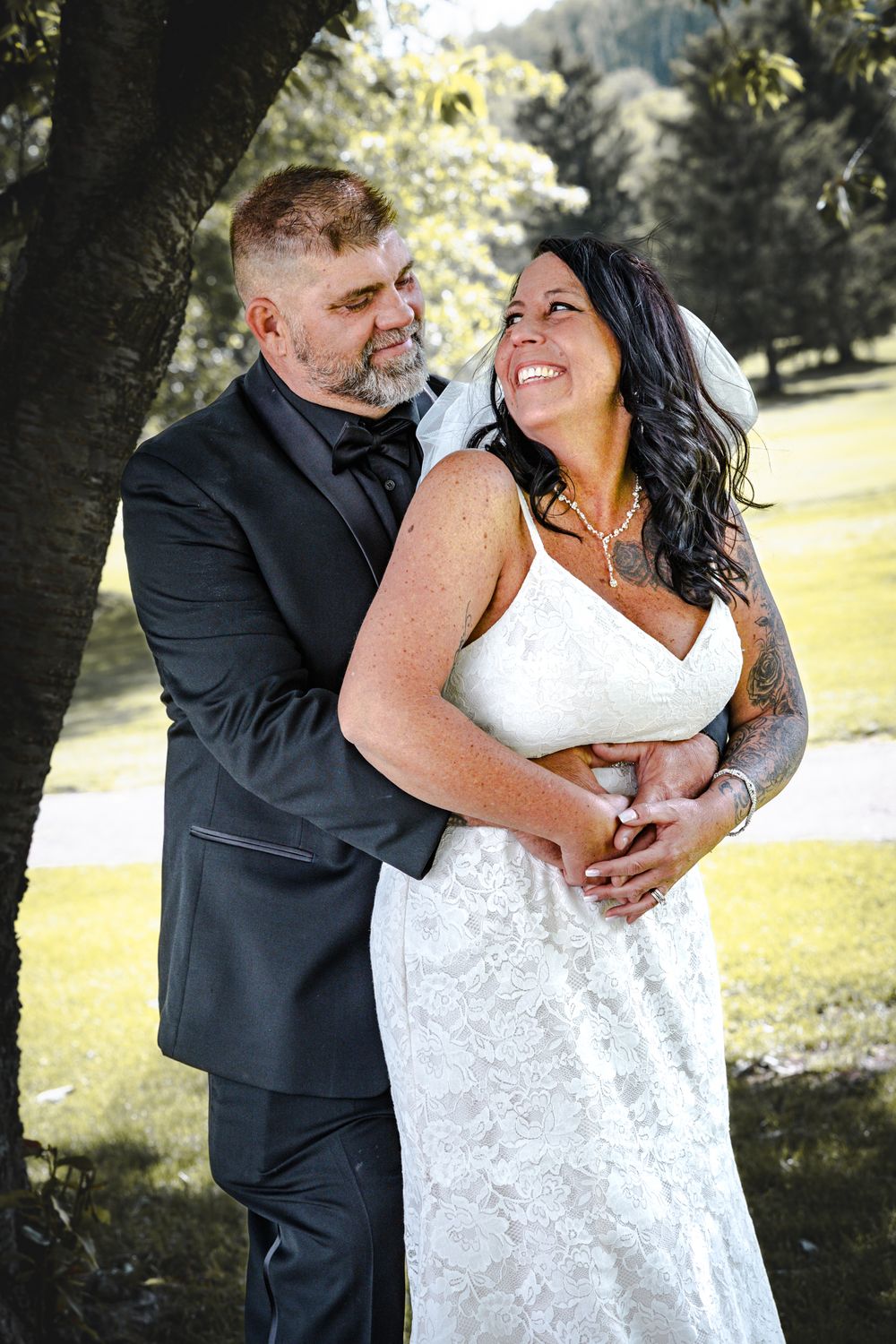A loving couple embraces under a tree on their wedding day, dressed in formal black suit and white gown.