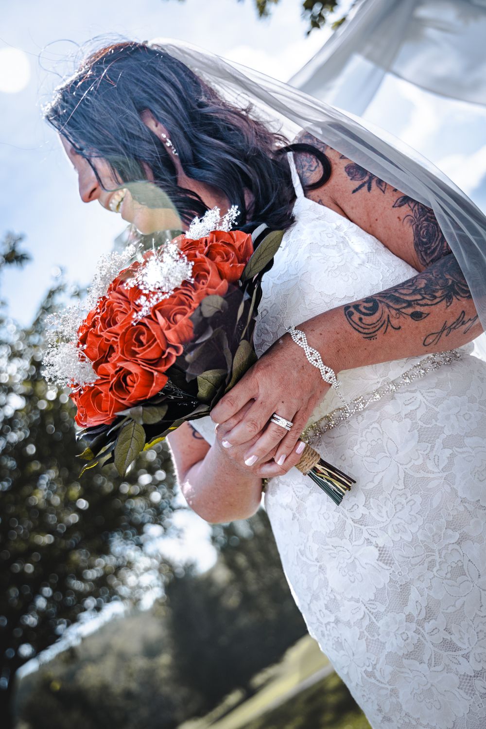 Close-up shot of white lace wedding dress and red rose bouquet against outdoor backdrop.