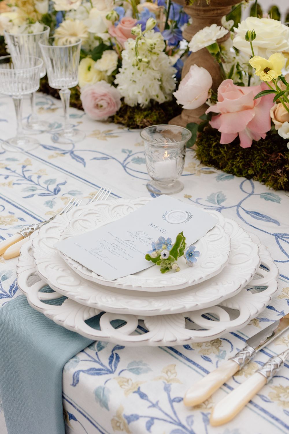 Elegant table setting with white plates, blue floral tablecloth, and pastel flower centerpiece with roses and hydrangeas.