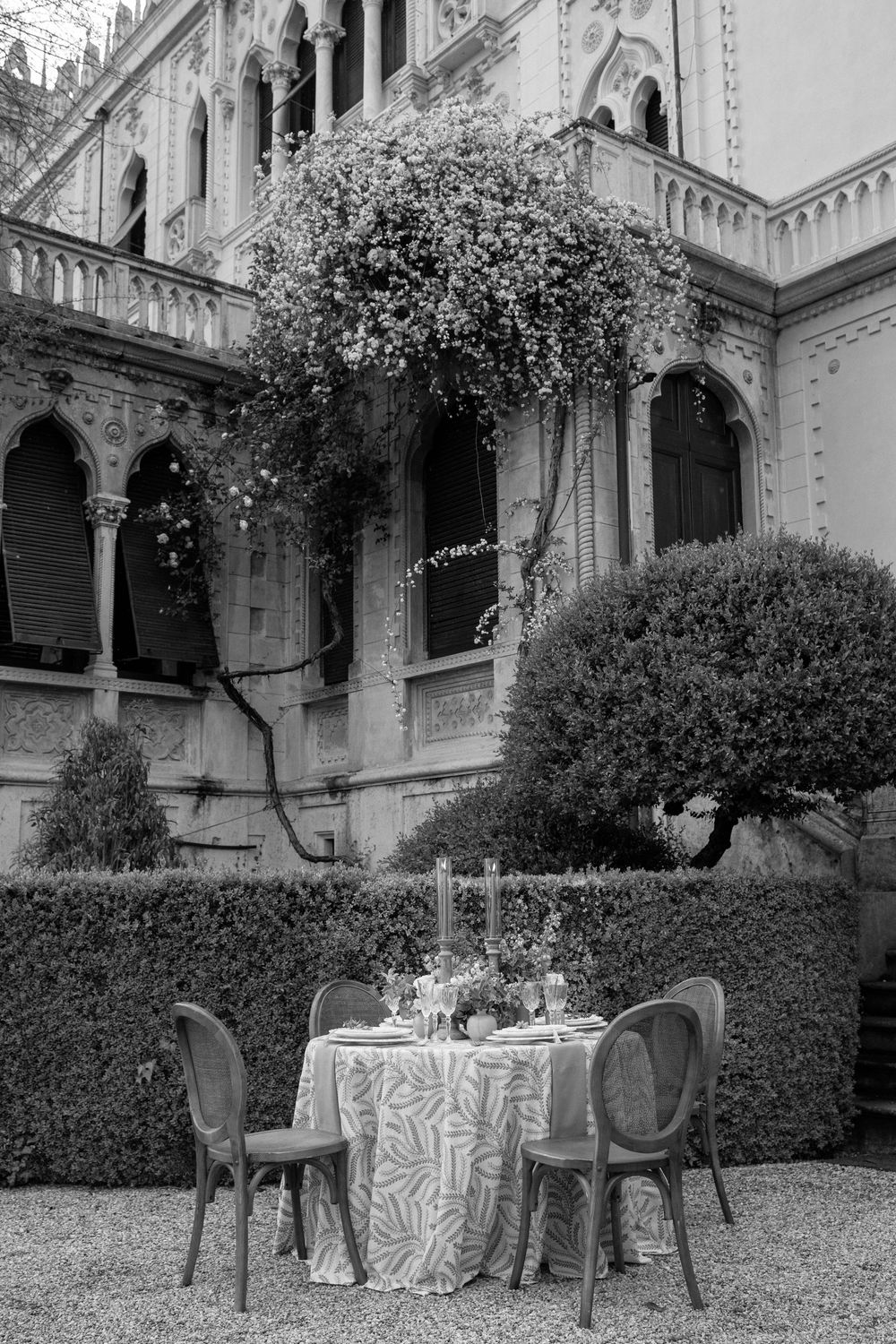 Five connected outdoor dining tables with elegant place settings arranged in a garden courtyard of a grand historic building.