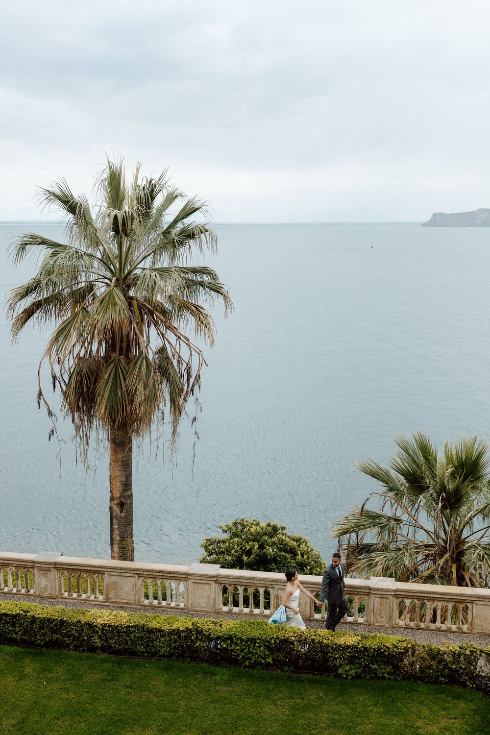A palm tree overlooks a misty oceanfront terrace with stone balustrades and manicured lawn at a coastal estate. Destination wedding Europe