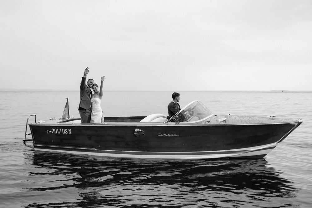 A vintage black and white photograph shows a classic wooden motorboat cruising across calm waters on a misty day.