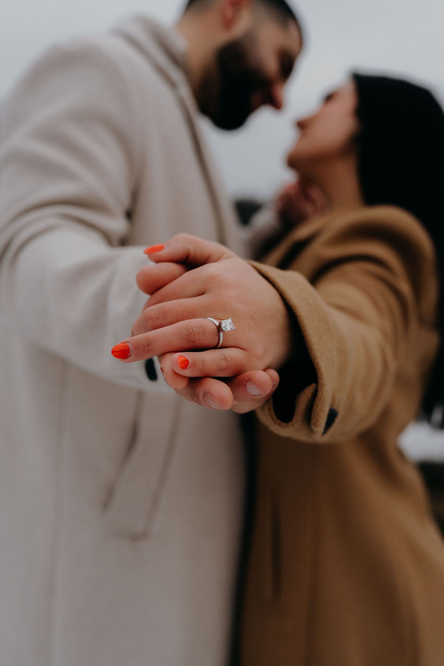 A romantic engagement photo showing clasped hands with a diamond ring and red nail polish.