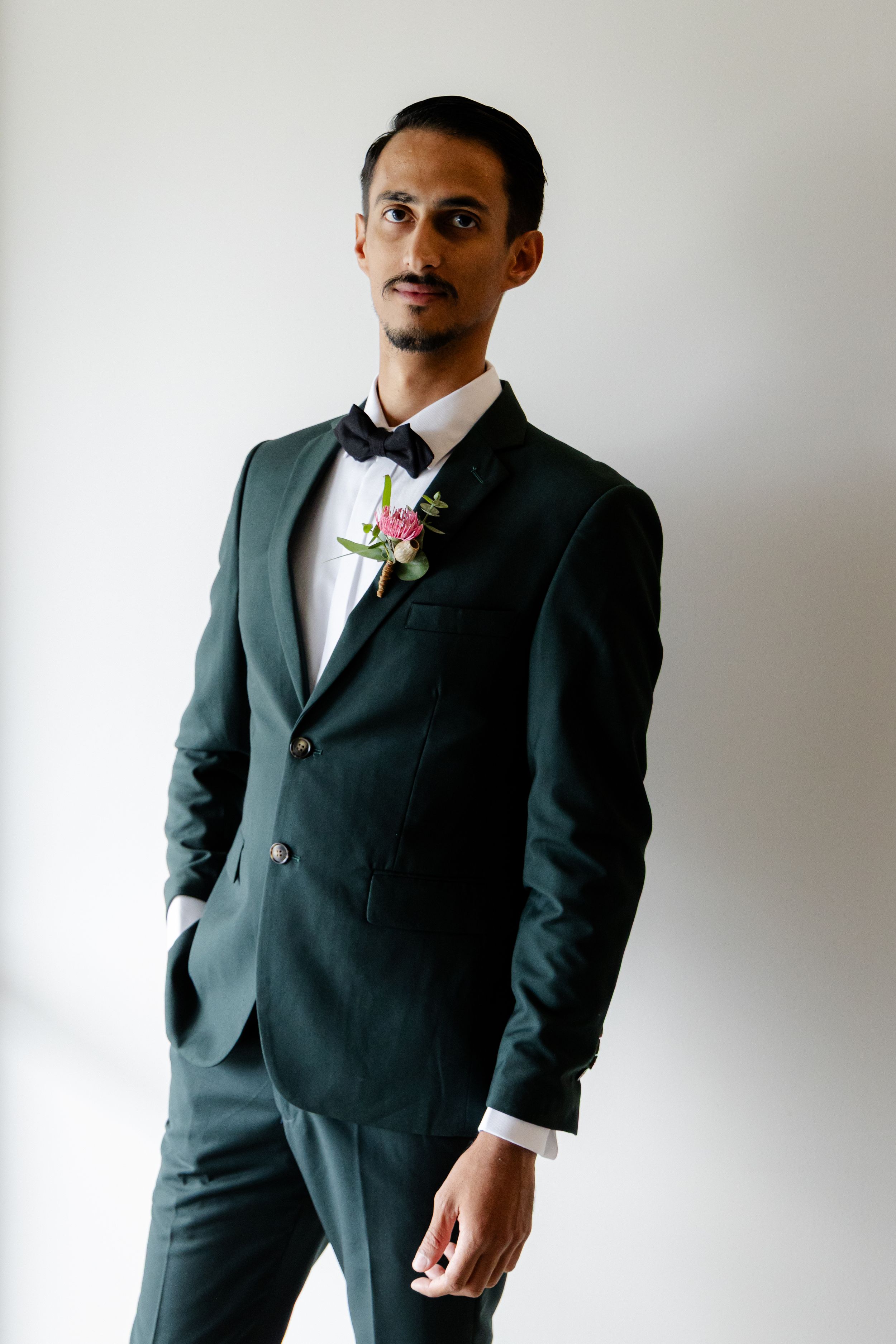 A groom poses in an elegant dark green suit with a pink boutonniere against a white backdrop.