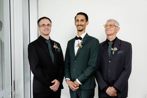 A line of men in dark formal suits and boutonnieres stand together against a white wall at an elegant event.
