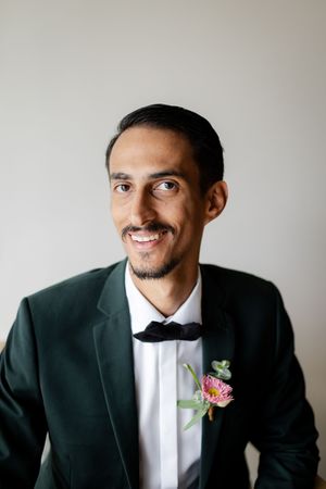 Groom in black tuxedo and white bow tie poses while wearing a pink flower boutonniere.