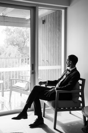 Black and white photo of someone sitting in a chair by a window in formal attire.