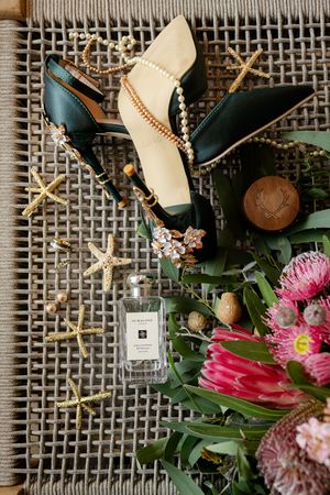 Wedding details flatlay with green heels, pearls, perfume bottle and pink protea flowers on textured gray surface.