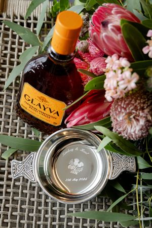 A bottle of liquor alongside pink protea flowers and greenery with an engraved silver quaich on a woven surface.
