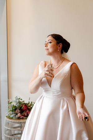 A person in a classic white wedding dress holds a perfume bottle near a window with soft natural light.
